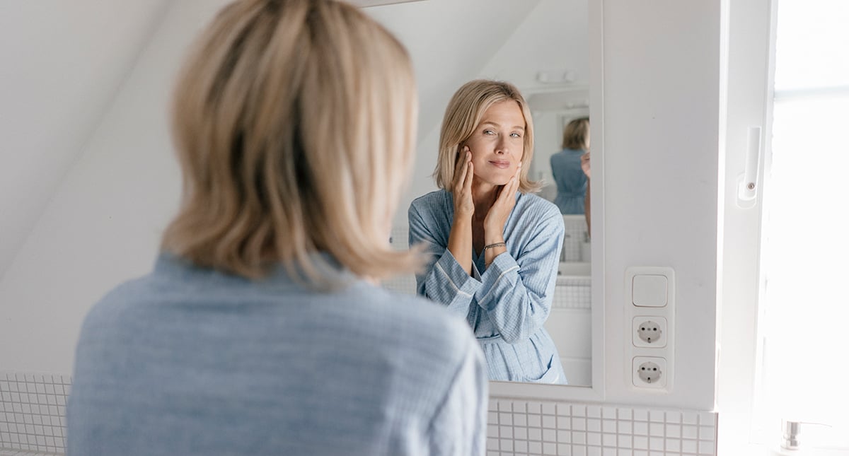 Medelålders kvinna står framför spegeln och studerar sitt ansikte.
