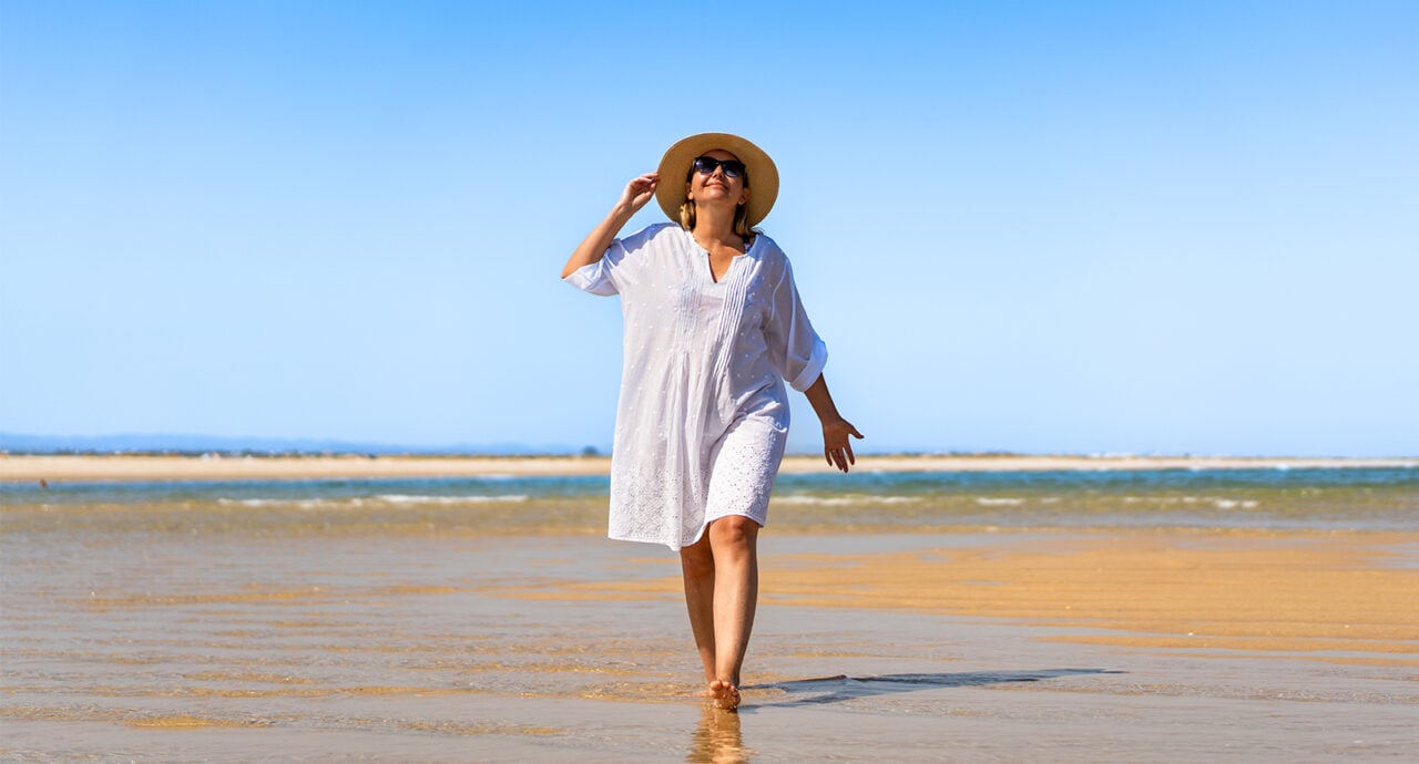 Kvinna i vit klänning och solhatt går på stranden i solskenet.