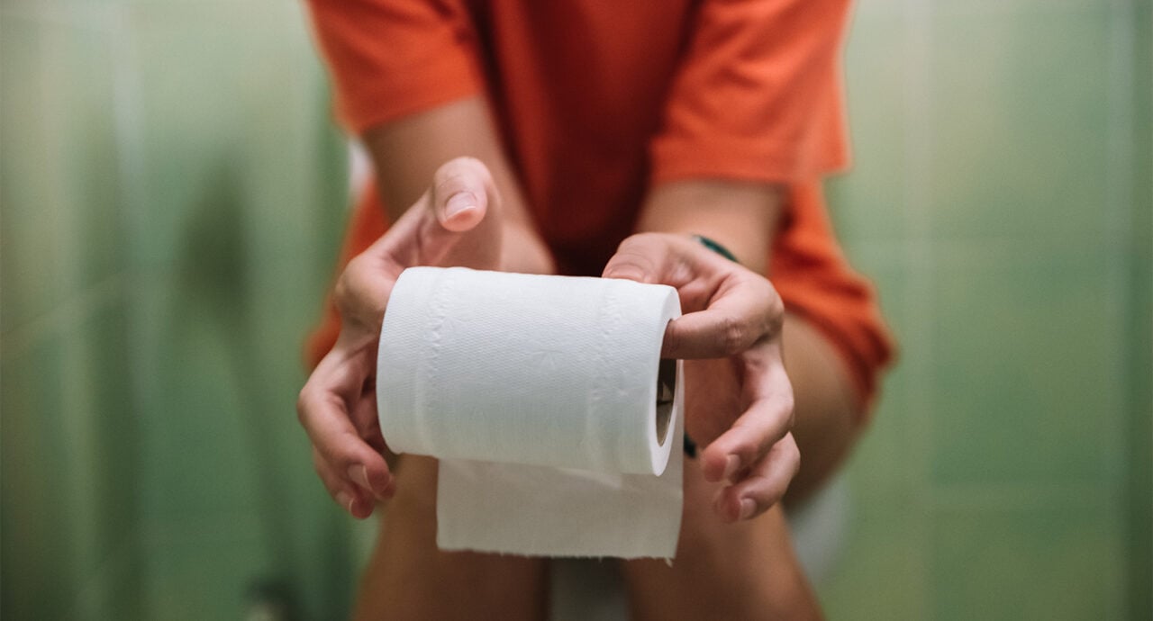 Kvinna sitter på toaletten, håller i en rulle toapapper.